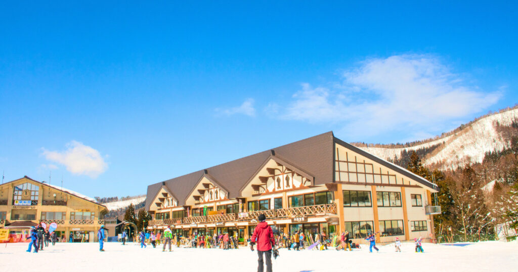 スキー場ロッジの周辺に、スキーを楽しむスキー客の画像。空は青く、スキー日和。